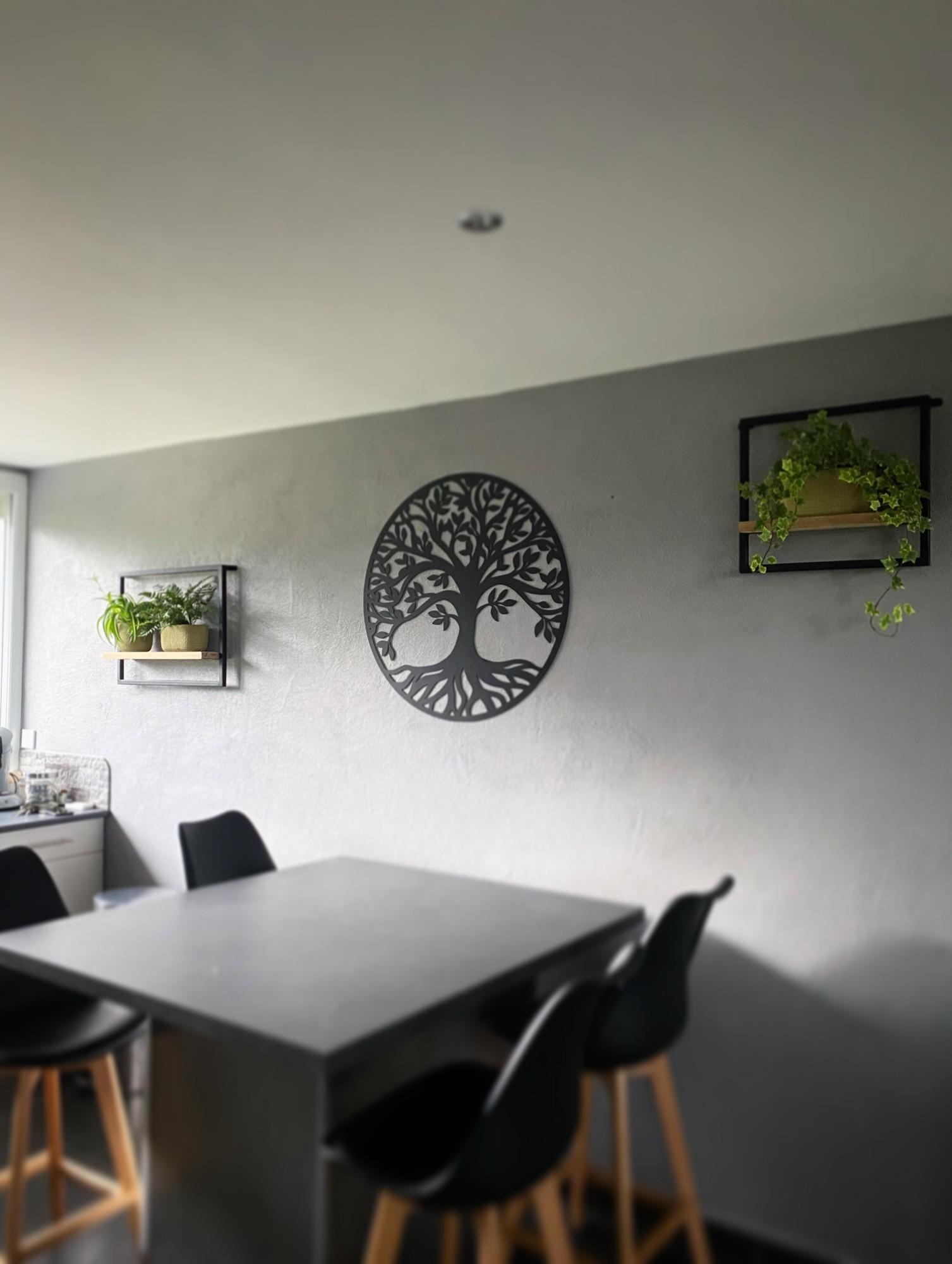 Arbre de vie circulaire en acier noir découpé au laser, accroché sur un mur gris au-dessus d’une table haute de cuisine, entre deux étagères garnies de plantes.