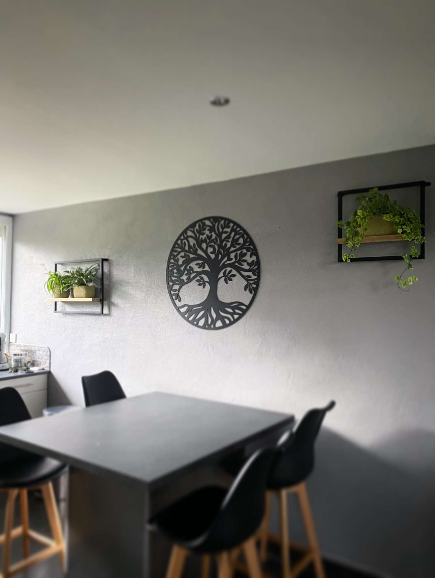 Arbre de vie circulaire en acier noir découpé au laser, accroché sur un mur gris au-dessus d’une table haute de cuisine, entre deux étagères garnies de plantes.