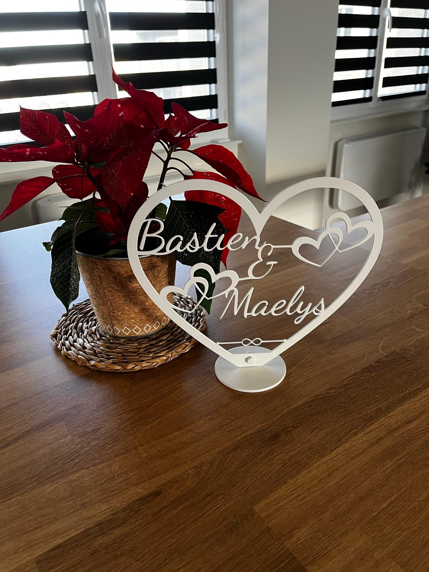 Cœur en acier blanc découpé au laser, portant les prénoms Bastien et Maelys entrelacés, posé sur son socle sur une table en bois, à côté d’une plante.