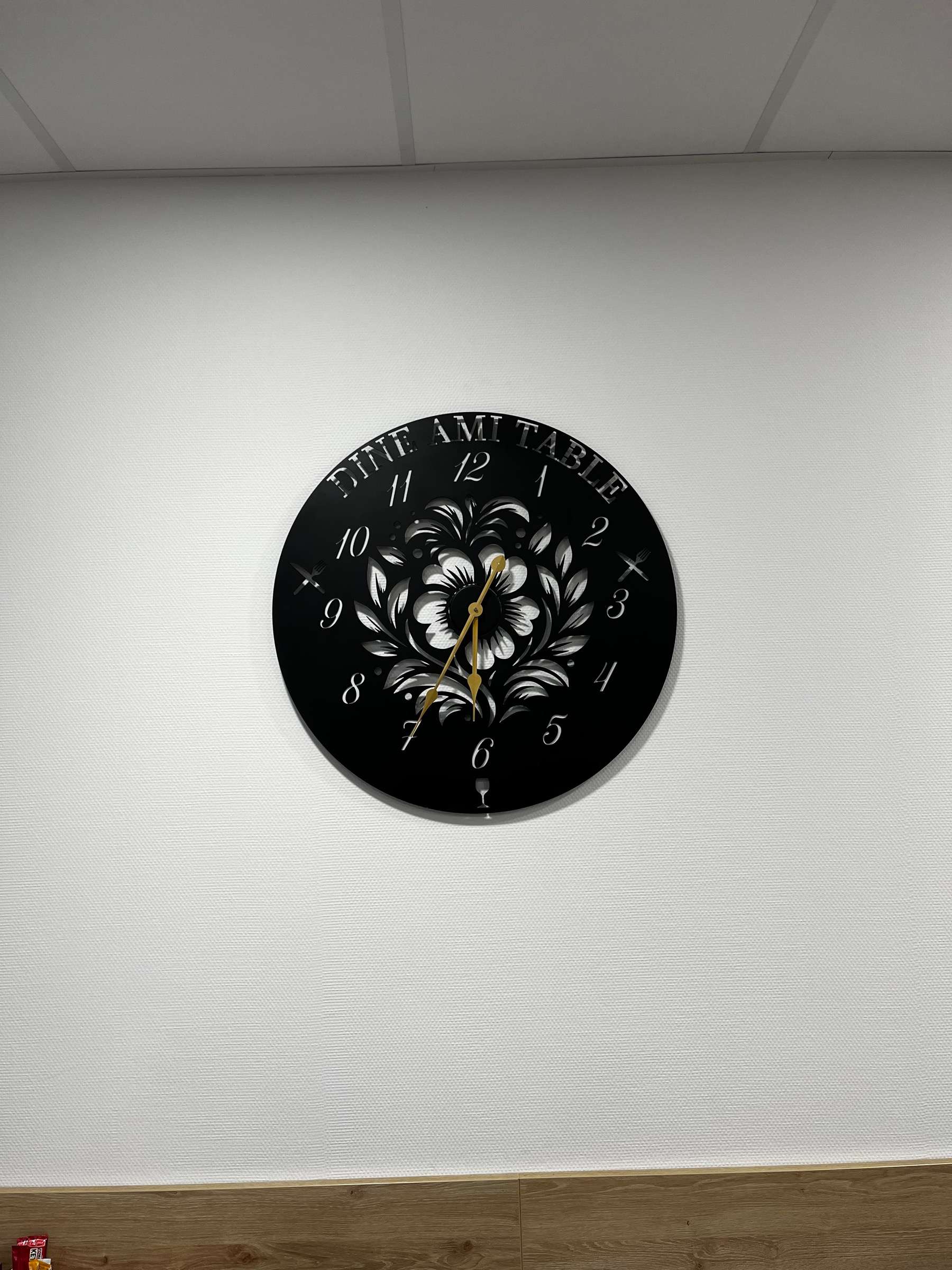 Grande horloge ronde en acier noir découpé au laser, avec l’inscription « Dine Ami Table », des chiffres ajourés, un motif floral au centre, des couverts et un verre de vin découpés, et des aiguilles dorées, fixée sur un mur blanc.