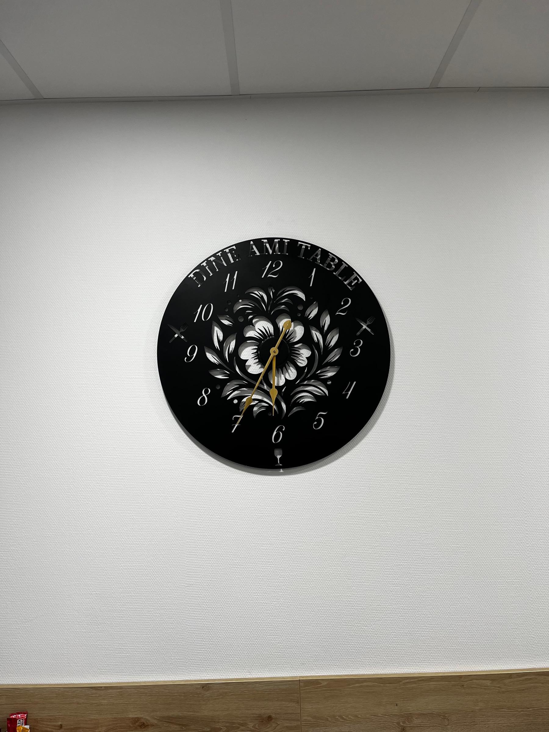 Grande horloge ronde en acier noir découpé au laser, avec l’inscription « Dine Ami Table », des chiffres ajourés, un motif floral au centre, des couverts et un verre de vin découpés, et des aiguilles dorées, fixée sur un mur blanc.